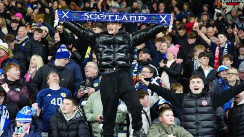 A crowd of Scotland fans celebrating in the stands at the match, with Denmark, which secured the Scotland team's place at the 2026 World Cup. A young boy can be seen in the centre of the frame, standing on the railings and holding up a Scotland scarf.