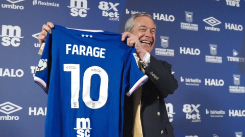 Nigel Farage poses with a blue Ipswich Town home shirt with 'FARAGE 10' on the back. He is standing in front of a blue advertising wall with various sponsors' logos on it. He is looking away from the camera and smiling.