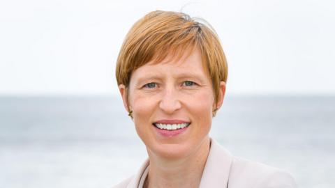 Clare Morley. She has short reddish brown hair and blue eyes. She is wearing a white blazer and is smiling at the camera.