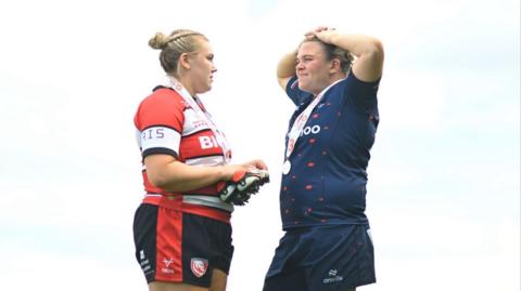 Mackenzie Carson on the left, wearing a black, white and red uniform. She is facing her partner Sarah Bern, who is wearing a blue uniform.