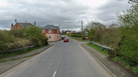Image of a rural road with residential properties on both sides and hedges of varying heights. Red and silver cars are visible in the distance, driving away from the camera, with a darker vehicle parked by the roadside.