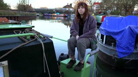 A woman is perched on the handrail of a houseboat on the River Lea