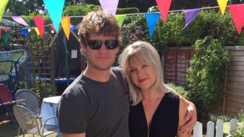 A man and a woman stand next to each other. The man, wearing a grey t-shirt, has his arms around the woman, wearing a black dress. They are in a garden with multi-coloured bunting above their heads.