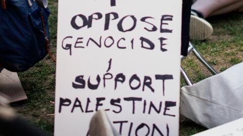 hand written poster with the words "I oppose Genocide I support Palestine Action" propped up by a person's feet on the grass. Lots of people nearby are seated on the grass. 