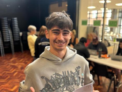 Boy smiling at the camera. Wearing a grey hoodie, moustache, brown hair and thick eyebrows.