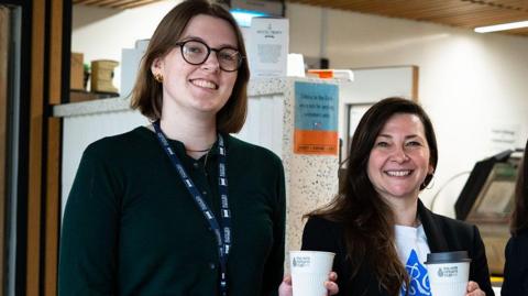 Ripley Hill-Nixon and Jane Martin. Ripley has short brunette hair and is wearing a dark green long sleeved top with dark trousers. She also has glasses. Jane has long brunette hair and is wearing a white t-shirt with a blue graphic, a black blazer and blue jeans. They are smiling and standing side by side, holding white coffee cups. 