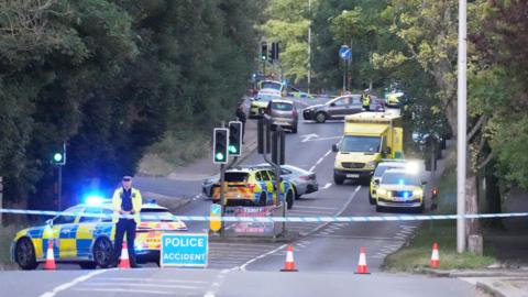 Four police cars and one ambulance are on a road. There is one police officer standing behind a cordon and five orange cones. There is also a blue and white sign, with the words "police accident".