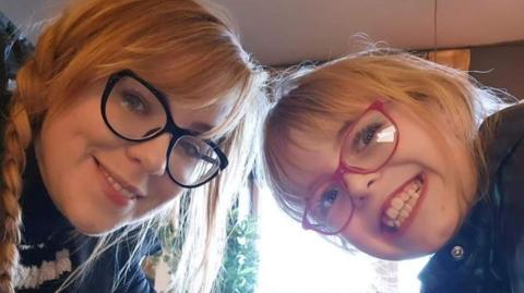 A picture of a mother with blonde hair beside her daughter. They have their heads tilted in towards each other both smiling. The mother is wearing black glasses and the nine year old is wearing pink glasses. 