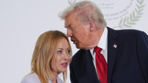 Donald Trump man on the right — dark suit, crisp white shirt, bold red tie — leans in with intent, his posture angled toward Italian PM Meloni. His expression is focused, almost as if he’s delivering something important quietly. Meloni, on the left, has light‑coloured hair and a pale outfit, is turned toward Trump, listening closely. Their body language is open but concentrated, suggesting the exchange is private, serious, or strategic rather than casual. Behind them, a large circular emblem reads SUMMIT FOR PEACE, placing the interaction in a diplomatic or political setting. 