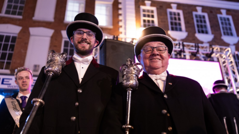 Two men wearing Victorian-era black suits and black top hats are pictured holding metal staffs with a royal crown at the top. 