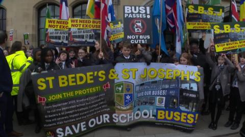 A group of schoolchildren holding posters and placards saying 'Save Our School Buses'