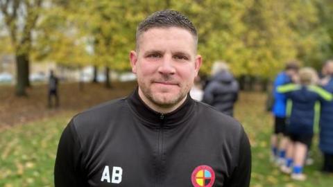 A man wearing a black training jacket stood in front of a green space and people in the background