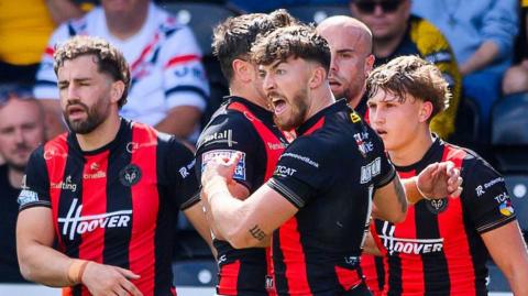 Warrington's Matty Ashton, in red and black stripes, shouts in celebration of his try as team-mates gather round to congratulate him.