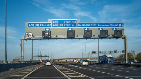 A motorway with six lanes - two are going to the left and the others are heading straight. There is a large gantry with road signs that are blue. One reads "the north west, birmingham north and west, m6"