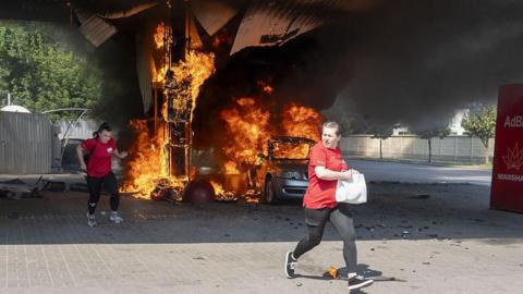 Two women in red tops run as flames rise from a car and fuel dispenser following a drone attack by Russia on a petrol station near the city centre of Kharkiv on 3 July