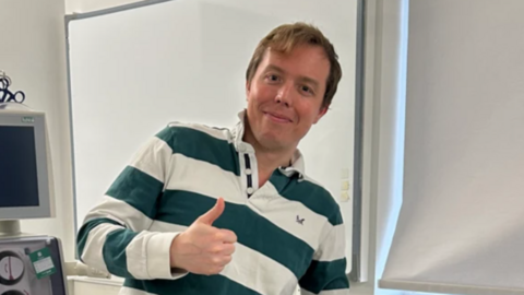 A man with short ginger hair and a polo top with green and white horizontal stripes with one thumb in the air and some medical equipment alongside him in a white room