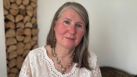 Deirdre McCanney has a neutral expression on her face as she sits in her front room. Behind her is a wood feature but it is blurred. Deirdre has shoulder-length  light brown and greying hair. She is wearing a white lace top with some eyelets. 