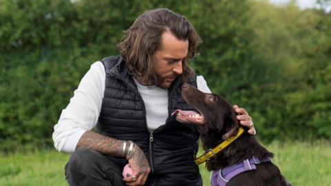 Pete Wicks, wearing a black gilet, white long-sleeved shirt and dark trousers, crouches down to stroke a chocolate-brown Labrador who gazes up at him with its tongue lolling out. The dog has a yellow collar. Pete has tattoos on his hand and arm. He holds a pink and black small ball. They are outside on a field.