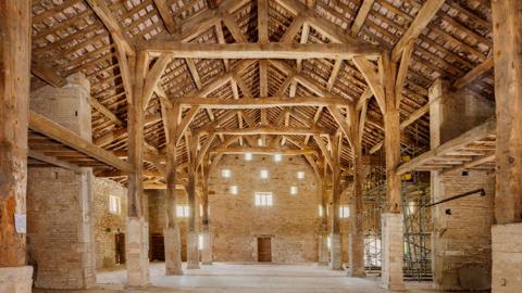 An image showing the barn's timber and stone interior. The colours are soft browns and show a high vaulted timber square frame attached to the roof trusses and showing the underside of the roof. There are eight wood pillars with stone bottoms supporting it, a stone floor and an arched wall is at the far end with seven small windows and a wooden door.