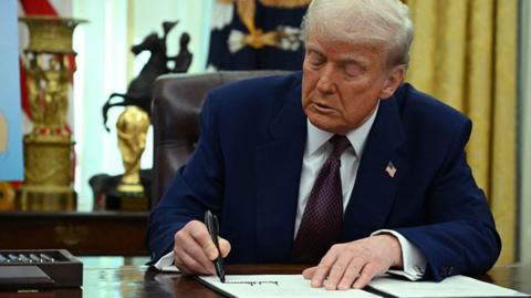 Donald Trump in the Oval Office of the White House signing a document 
