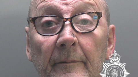 James Hughes police mugshot. He has stubble and is wearing spectacles