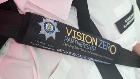 Image shows a police officer wearing the black seatbelt cover, which says on it in blue and yellow writing: "VISION ZERO PARTNERSHIP, Delivering Safer Roads for Cambridgeshire & Peterborough. SECURE PEGASUS SCHEME PIN INSIDE". There is the Cambridgeshire Police logo on it.