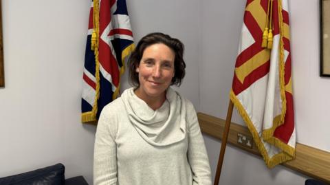 A lady in a white jumper stood infront of union flag and a Guernsey flag
