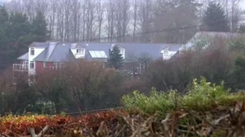 Holmesley Care 鶹Լ, pictured from a distance. The home is a multi-storey building with a red brick exterior surrounded by trees.