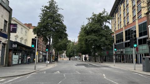 A general view of Upper Parliament Street Nottingham, taken from the middle of the road, in front a pedestrian crossing showing a patch of darker Tarmac in the road, where Severn Trent works were recently completed. Two restaurants can be seen on either side of the street and there are a number of trees lining the street in the background 