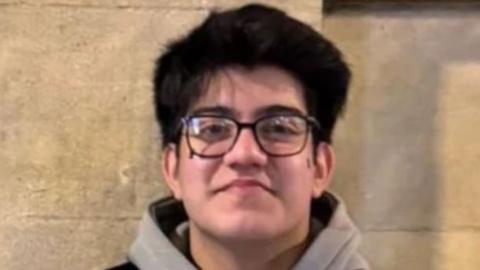 Taha has medium length black her and black rim glasses. He has a grey hoodie and a black gilet