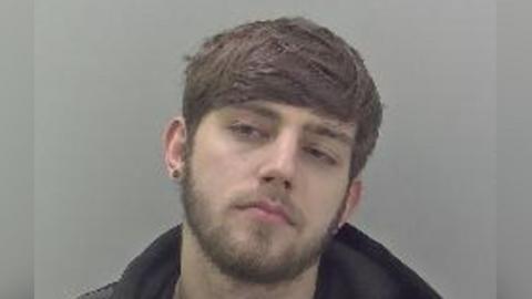 A police photograph of paedophile Scott Chapman. He is waring an earring, has brown hair and a beard. He is refusing to look straight into the camera, tilting his head. He is also wearing a dark jumper and hoodie. 