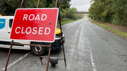 A red Road Closed sign on a brown stand, put across the middle of the carriageway. Trees line the road. A white van is parked behind the sign.