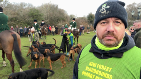A picture of Will Day wearing a woolly hat that says "New Forest Hounds" and a green bib that says "New Forest Bloodhounds Quarry Runs". 