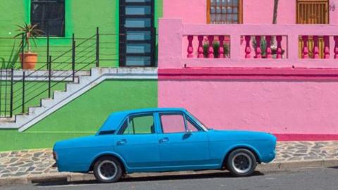 A bright blue vintage-style car is parked on a quiet street in front of two vividly painted buildings. The left building is lime green with a black‑framed door and a white staircase bordered by a metal railing. A small potted plant sits near the top of the stairs. The right building is painted pink with a decorative balcony, wooden-framed windows and a smooth plastered wall. The bold blocks of green, pink and blue create a striking, colourful scene.