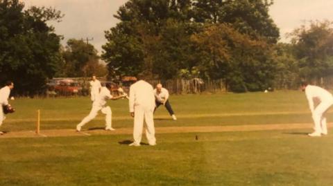 A batsman plays a forward defensive shot watched by three fielders, a wicketkeeper and an umpire, during a cricket match in Shepreth, Cambridgeshire.