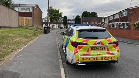 A picture of a police car parked on a street. Two-storey maisonettes can be seen in the background. There is a grass verge lining the pavement.