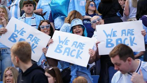 Coventry City fans dressed in Sky Blue in the stands of the Coventry Building Society Arena. the image centres on three fans holding up We Are Back signs. The woman in the centre has sky blue dyed hair and a blue bucket hat. On her right is a man with heart-shaped sky blue glasses and on her left a woman in the same glasses with long dark hair.