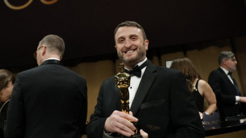 Mike Hill, wearing a black tuxedo, smiles as he hold an Oscar backstage at the Oscars ceremony