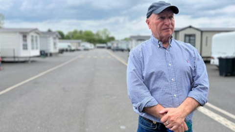 Billy Welch wears a baseball cap, stands with clasped hands with a row of caravans behind him