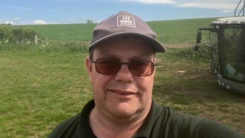 A selfie of a male farmer who is standing in a field. His tractor is next to him. He is wearing a blue hat and a green top. There is a green field behind him. 