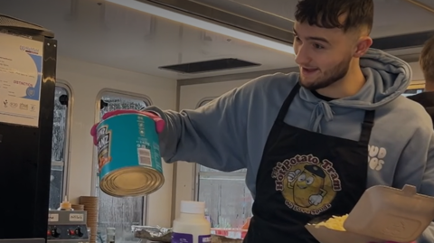 Spud Brother Harley Nelson prepares a baked potato at the counter of the Spud Brothers' van. He is holding a baked potato in a styrofoam tray in one hand and a catering-sized tin of baked beans