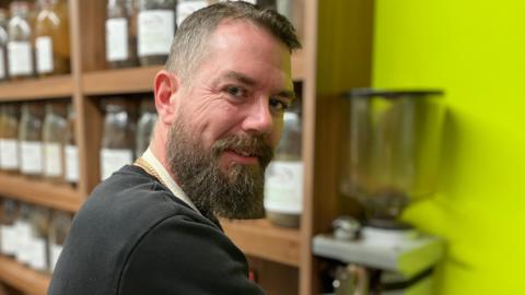 Nigel Lyons has a brown beard and short brown hair and is wearing a black jumper is pictured in his coffee shop in front of jars of coffee beans