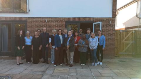 A group of 16 people posing for the camera outside the new house. The 10 women and six men are standing in front of patio doors of the brick building on a stone patio.