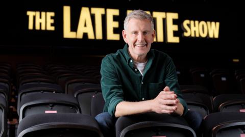 Paddy Kielty, wearing a dark green shirt with a white t-shirt underneath, and wearing a pair of dark trousers. He has short grey/blonde hair. He is sitting in the audience seats, in the middle of an empty studio with the words 'The Late Late show' in the background.