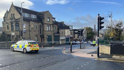 A police car is parked across the junction of a road that has been taped off with blue and white crime scene tape.