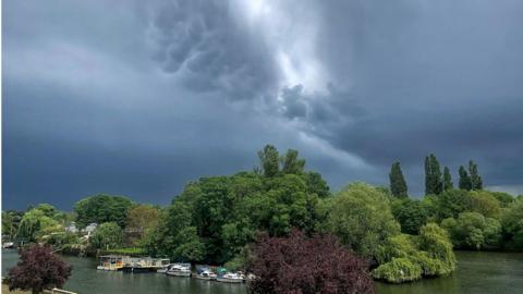Dark cloud with a crack of light down the centre, above a treeline on a river front