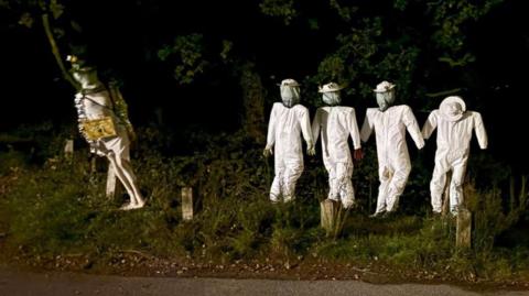 Five scarecrows on a grass verge in the dark. One stood alone on the left is wearing white, a hat and a sign depicting a bee. The other four, to the right, are wearing white beekeeping uniforms and look similar.
