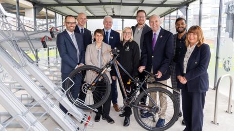 Noel Lavery (WMRE), Andy Thrupp (WMCA), Beccy Marston (TfWM), Rob Goode (WMR), Claire Williams (TfWM), Cllr Chris Burden, Hugh Porter MBE, Asish Chandragiri (WMR), Teresa Woodward (WMR) stand behind a bike in the new hub. They are all smartly dressed in suits, or jackets and trousers and smiling at the camera.