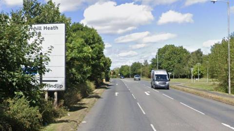 A road with trees down either side and a couple of cars travelling along it and a white road sign on the left hand side