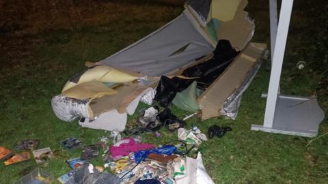 Various items of waste seen scattered on a grass area. There appears to be a sofa as well as a mirror and several food packets.